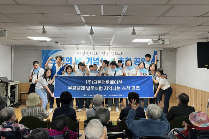 요양원 실내 무대에서 '(주)크린팩토메이션 우쿨렐레 별꽃처럼 지역나눔 초청 공연'이라고 적힌 파란색 현수막을 함께 들고 환하게 웃고 있는 연주단원들의 모습.