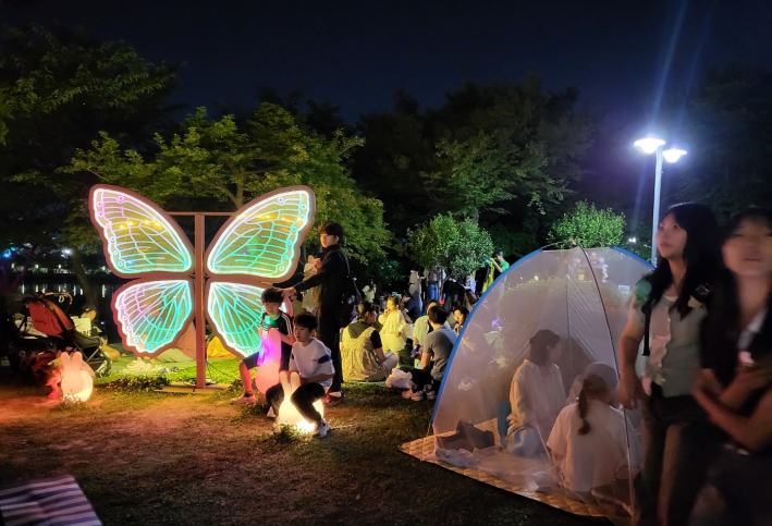 형형색색 나비 조형물 아래에서 사진을 찍는 아이들과, 모기장 안에서 벌레 걱정 없이 축제를 즐기는 가족들. 평화롭고 따뜻한 여름 밤의 아이디어가 돋보인다.