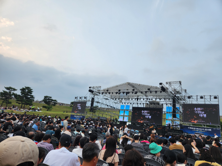 수백 명의 관객이 질서정연하게 착성해 공연 시작을 기다리고 있고, 무대 양 옆의 대형 스크린에는 '수원화성 헤리티지 콘서트 2025'와 '수원, 감각유산 여행'이라는 문구가 선명하게 보인다. 
