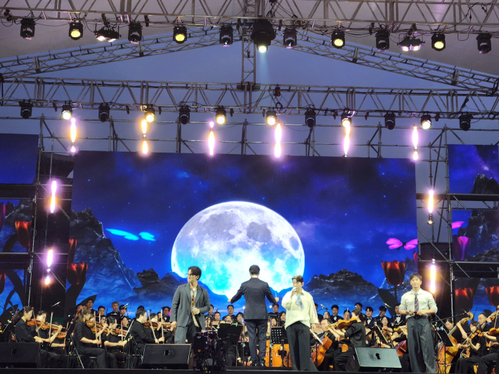 밤하늘과 보름달, 환상적인 조명 아래 오케스트라와 함께 여러 명의 출연자가 무대에 올라 합창과 특별 공연을 선보이고 있다. 