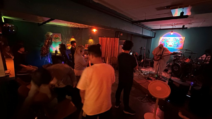 오른쪽 무대 위에는 밴드가 공연을 하고 있고, 무대 앞에 한명이 카메라를 들고 촬영하고 있다. 주위에는 좌측 출입구까지 꽉 찬 관객들이 저마다 방식으로 공연을 즐기고 있다.