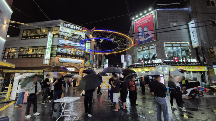 버스킹을 즐기며 통닭집 앞에 많은 시민들이 우산을 들고 서있다.