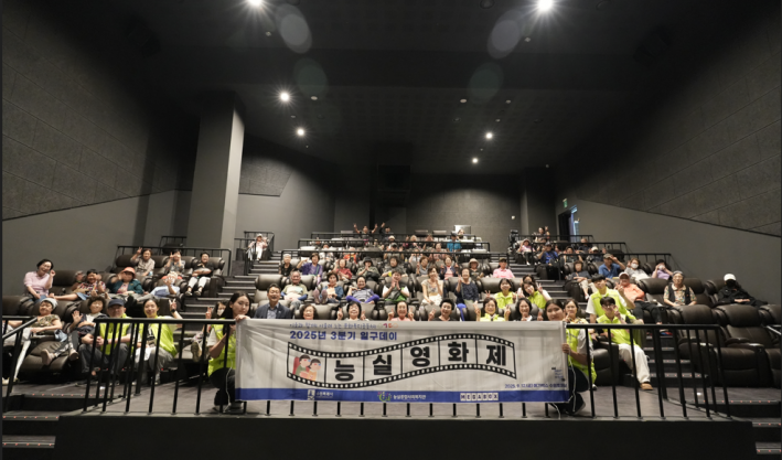 능실마을 주민들이 영화관 좌석에 앉아있고, 일구데이 활동단과 기획참여주민은 연두색 조끼를 입고 앞쪽에 앉아 현수막을 들고 단체 사진을 찍었다.