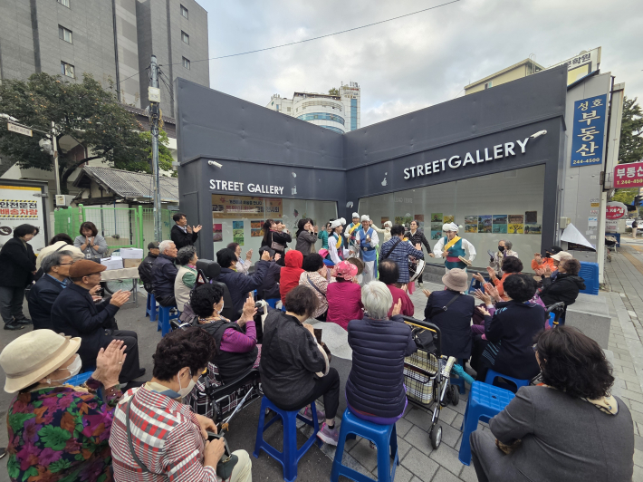 갤러리 앞에서는 전통 복장을 입은 공연팀이 사물놀이 를 선보이고, 참석자들은 박수로 호응하며 즐거운 분위기를 연출하고 있습니다.