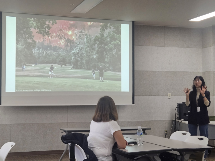 산불이 났음에도 그 아래서 한가롭게 골프를 치고 있는 사람들 사진을 놓고 환경강사가 환경의 심각성을 이야기하고 있다.