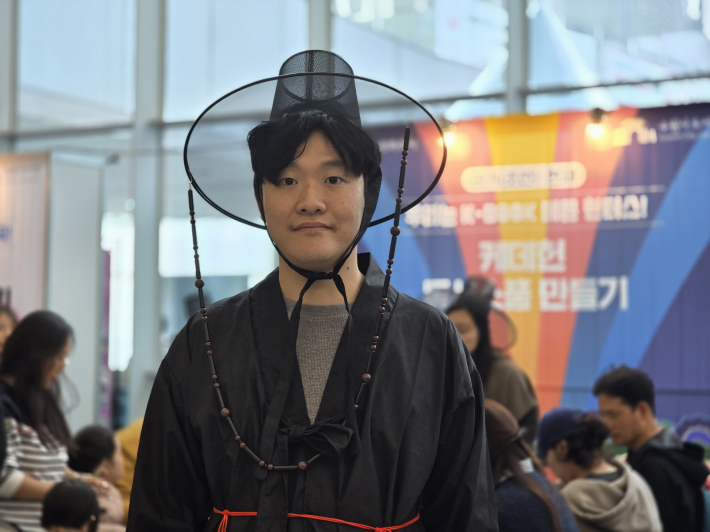 갓을 쓴 진행요원, '케데헌'의 수호자 콘셉트로 참여.  '우리는 K-BOOK 데몬 헌터스!' 부스의 진행요원이 전통 갓을 착용하고 시민들을 맞이하고 있다.