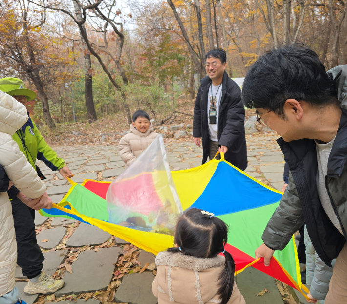 가족과 아이들이 함께 알록달록한 패러슈트를 잡고 낙엽 풍선을 높이 띄워 올리며 협동 놀이를 하는 장면.