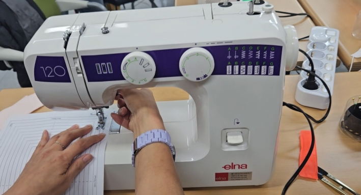 종이 위에 그려진 선을 따라 박음질 하는 재봉틀 사용법을 강의하고 있는 강사의 모습 