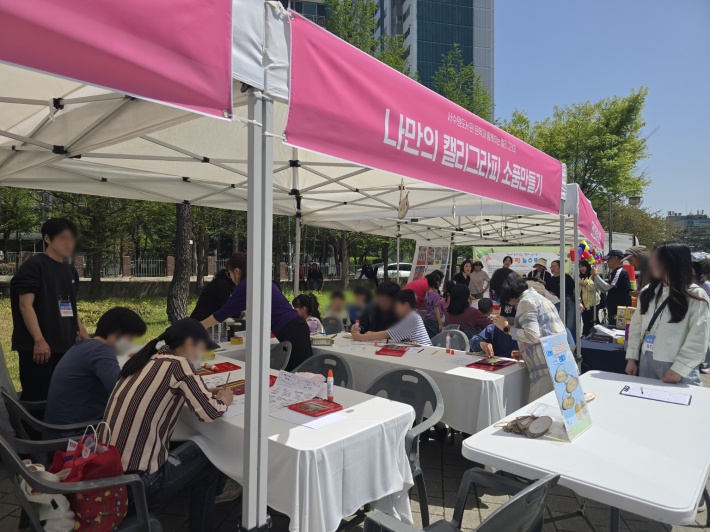 야외 체험부스에서 아이들과 가족들이 '나만의 캘리그래피 소품 만들기' 활동에 참여하며 즐거운 시간을 보내고 있다.