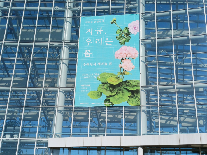 전시 온실 창, '지금 우리는 봄' 제라늄 홍보 글이 멀리서 보인다.