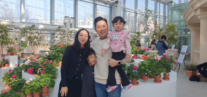 설 연휴를 맞아 수목원 온실을 찾은 4인 가족 관람객이 활짝 핀 제라늄을 배경으로 밝게 미소 지으며 기념사진을 찍고 있는 모습