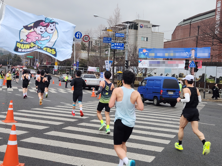 'RUN RUNNERS' 깃발이 휘날리는 가운데 참가자들이 성균관대역 인근 교차로를 지나고 있다. 시민들의 응원 속에 도심은 하루 동안 거대한 트랙이 되었다.