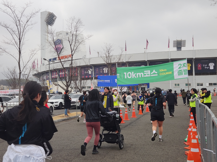 수원종합운동장 일대에서 열린 마라톤 대회에서 참가자들이 '10km 코스 출발' 구간을 지나 힘차게 달리고 있다. 가족 단위 참가자와 유모차 러너까지 함께해 시민 참여형 생활체육 축제의 분위기를 보여준다.