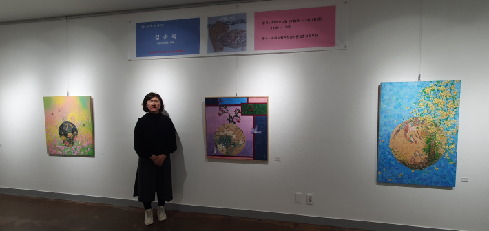 전시장에서 작품 앞에 선 김순옥 작가. 위로 전시 안내 현수막과 왼쪽과 오른 쪽으로 출품작들이 보인다.