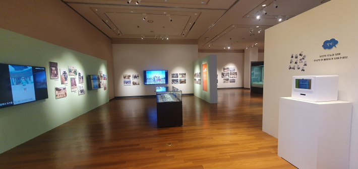 3부 전시 공간, 중앙에 길쭉한 유리 진열장이 있고, 벽면에는 모니터 영상과 시민들이 제공한 컬러 사진들이 걸려 있는 내부 모습.