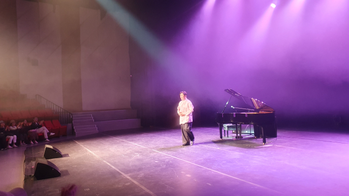 보라빛 무대 조명 아래 고영일이 그랜드 피아노 옆에 서서 관객과 소통하고 있다.