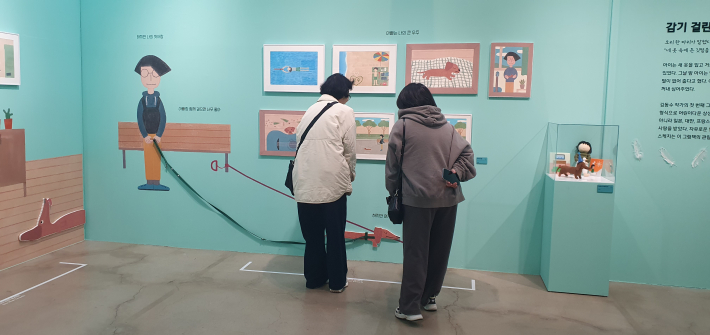관람객들이 김동수 작가의 그림책 원화와 벽화를 감상하고 있다. 민트색 전시장 벽면에는 아이와 아빠, 반려견 산책 장면이 이어진다.