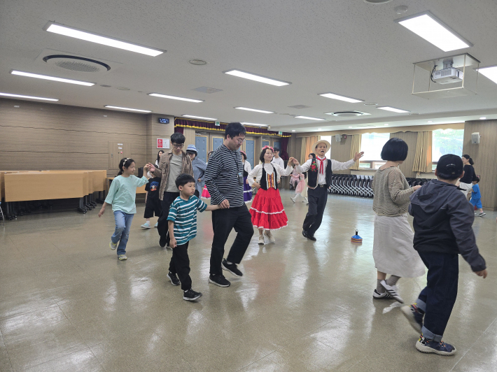 아이와 부모가 함께 발을 맞추며 포크댄스를 따라 하고 있다. 가족이 서로의 손을 잡고 움직이며 웃음과 교감을 나누는 모습이 인상적이다.