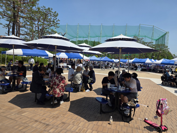 야외 광장 파라솔 아래 모여 앉아 바자회에서 구매한 시원한 음료와 간식을 나누며, 무더위를 피해 휴식을 취하고 있는 시민들의 모습.