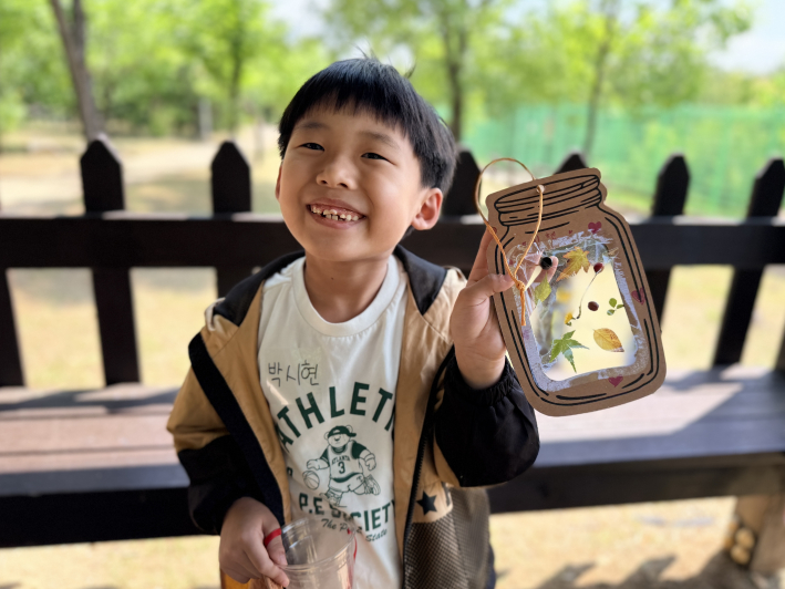 숲 체험 활동 후 자연물 작품을 들고 환하게 웃는 아이. 작은 잎과 꽃, 열매를 모아 만든 작품에는 아이가 직접 만난 봄의 흔적이 담겼다.
