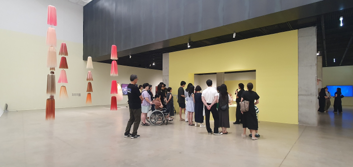 관람객들이 머리카락을 이용한 설치작품과 조각이 놓인 전시장에 모여 작품을 살펴보고 있다. 휠체어를 이용한 관람객을 비롯해 다양한 관람객이 전시를 함께 보고 있다.