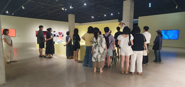 관람객들이 전시대 주변에 모여 조각 작품을 바라보고 있다. 오른쪽 벽면에서는 영상 작품도 상영되고 있다.