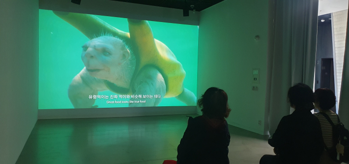 관람객들이 초록빛 영상이 상영되는 전시실에 앉아 작품을 감상하고 있다.